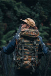 Person wearing a camouflage caribee rucksack and hat crossing a forest bridge