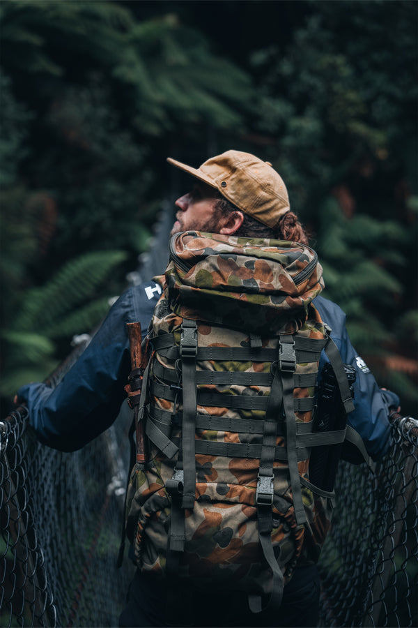 Person wearing a camouflage caribee rucksack and hat crossing a forest bridge