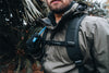 Man wearing a grey jacket with a hydration backpack in a forest setting