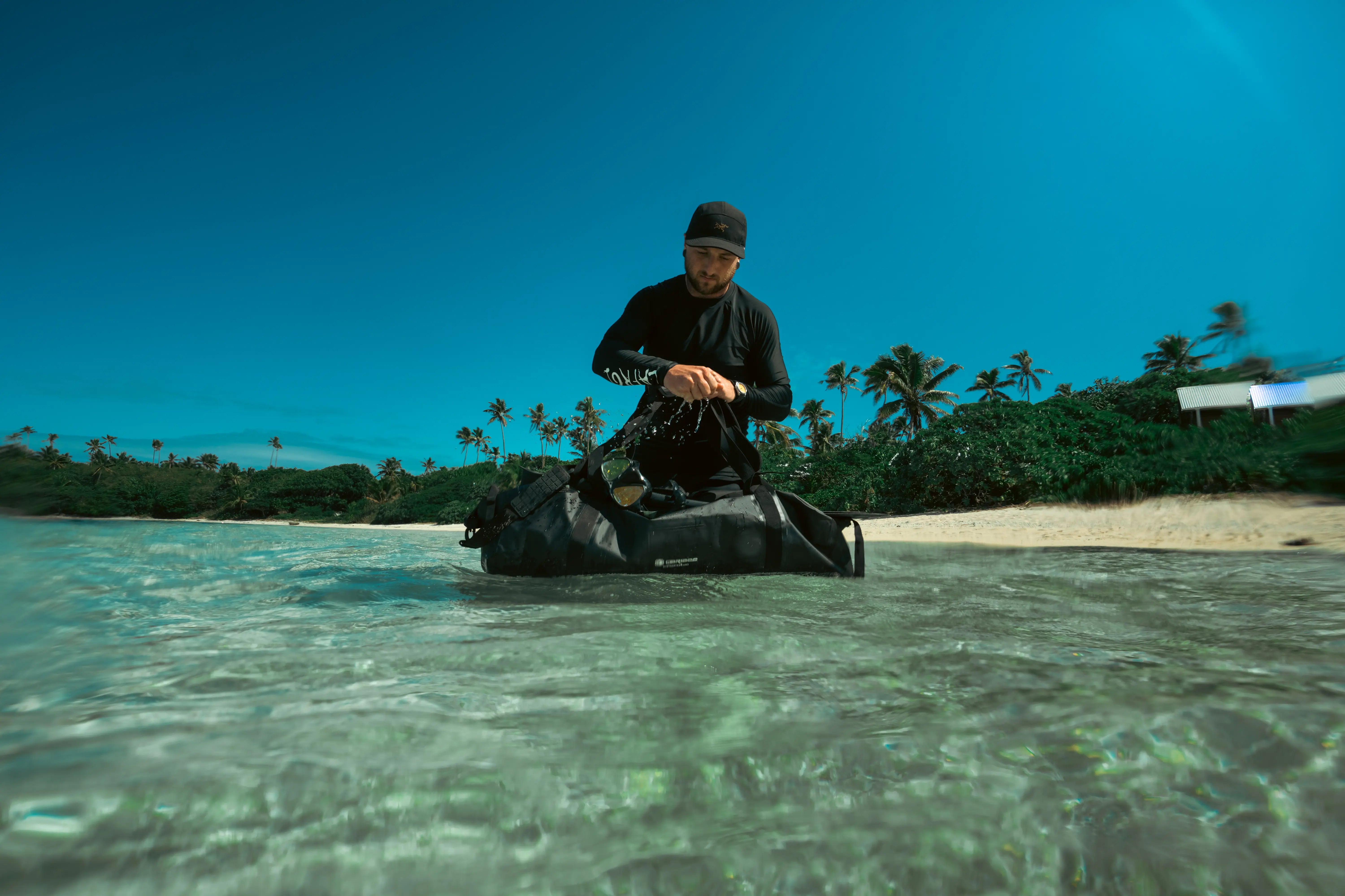 Person with Caribee water proof bag in shallow water near a beach with palm trees
