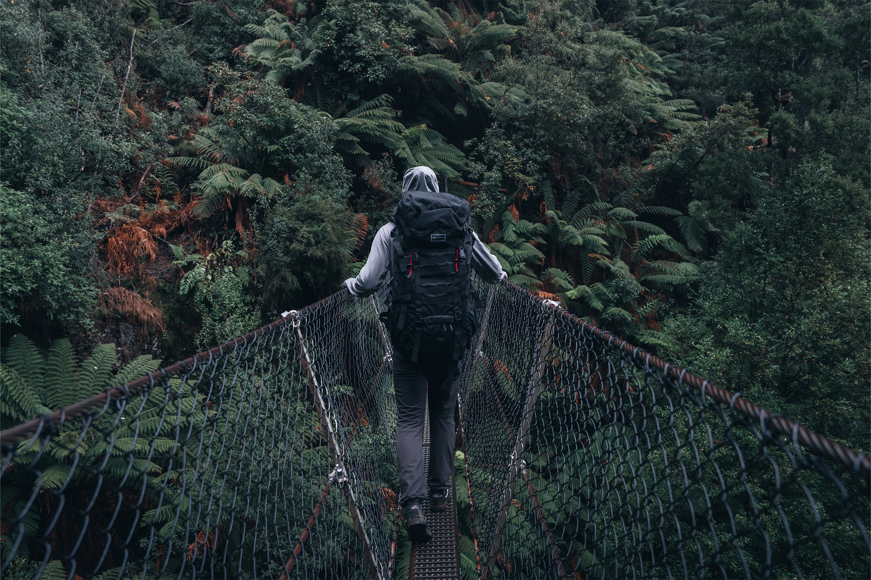 Person with a black Caribee Pulse rucksack crossing a suspension bridge in a forest