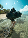 Person carrying a large black Caribee waterproof bag through shallow water on a beach.
