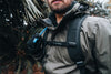Man wearing a grey jacket with a hydration backpack in a forest setting