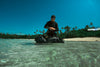 Person with Caribee water proof bag in shallow water near a beach with palm trees