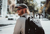 A man wearing a black baseball cap, sunglasses, a beige shirt, and a black Caribee Komodo backpack walking on a sunny city street.