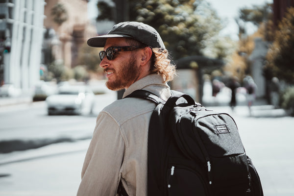 A man wearing a black baseball cap, sunglasses, a beige shirt, and a black Caribee Komodo backpack walking on a sunny city street.