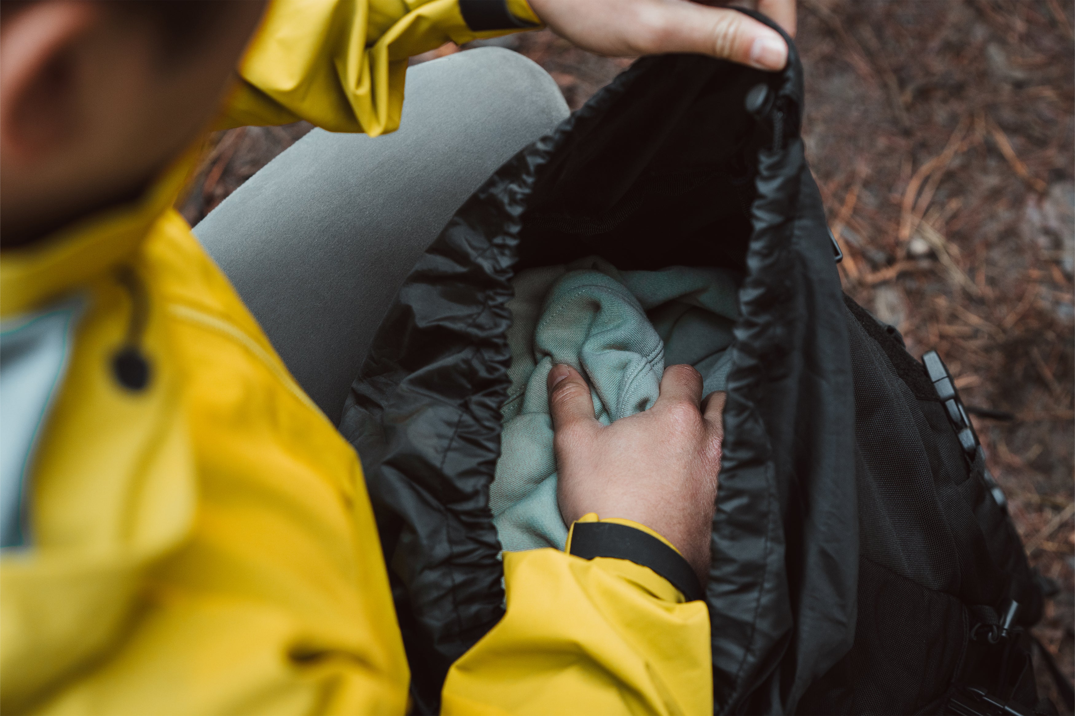 Person in yellow jacket packing a green jumper into a black Caribee rucksack outdoors.