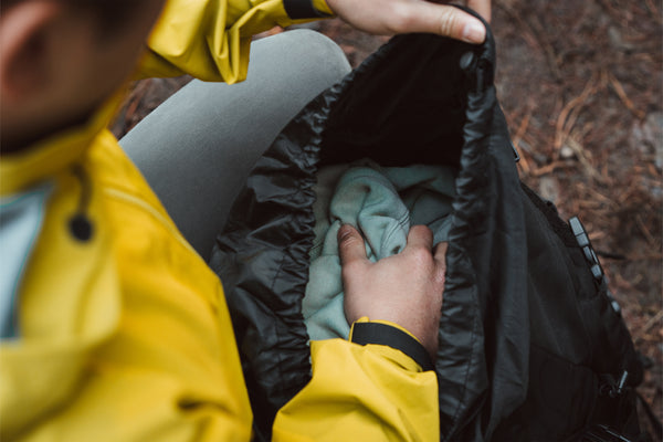 Person in yellow jacket packing a green jumper into a black Caribee M60 Phantom rucksack in the outdoors.