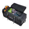 A black Caribee Scarecrow bag shown open, filled with gear including an orange and black helmet, a green bag, a gray cap, and blue jeans, on a white background.